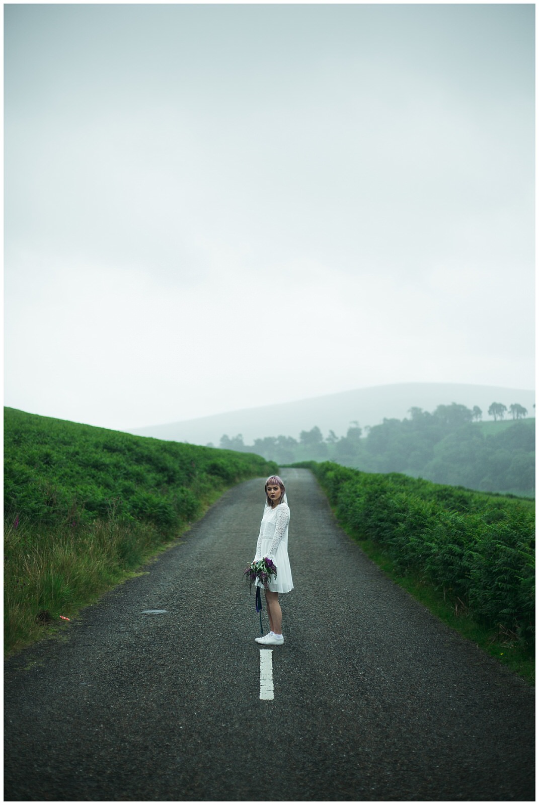 Bride in a short boho wedding dress in lace and satin stands on a deserted mountain road in Ireland